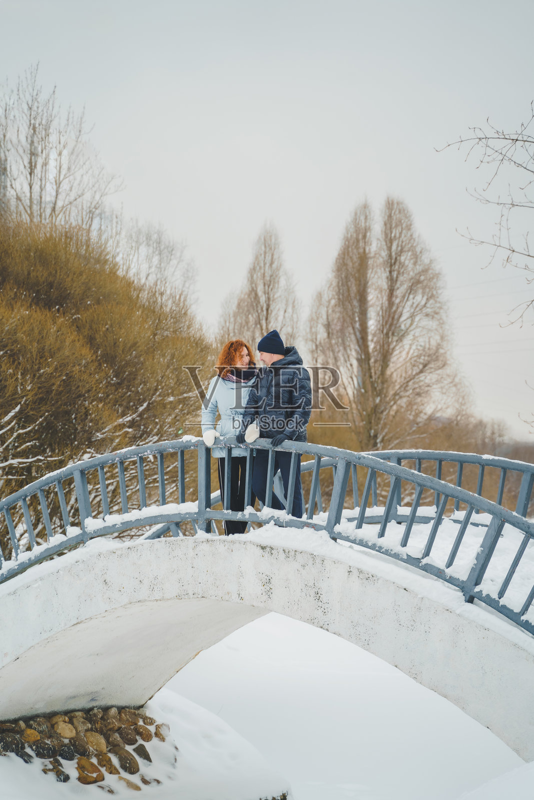 浪漫爱情故事。快乐的中年千禧一代情侣，冬季雪地公园里一位红发女子。浪漫约会，亲吻，拥抱。散步，享受乐趣。时尚的蓝色衣服，夹克，帽子。照片摄影图片