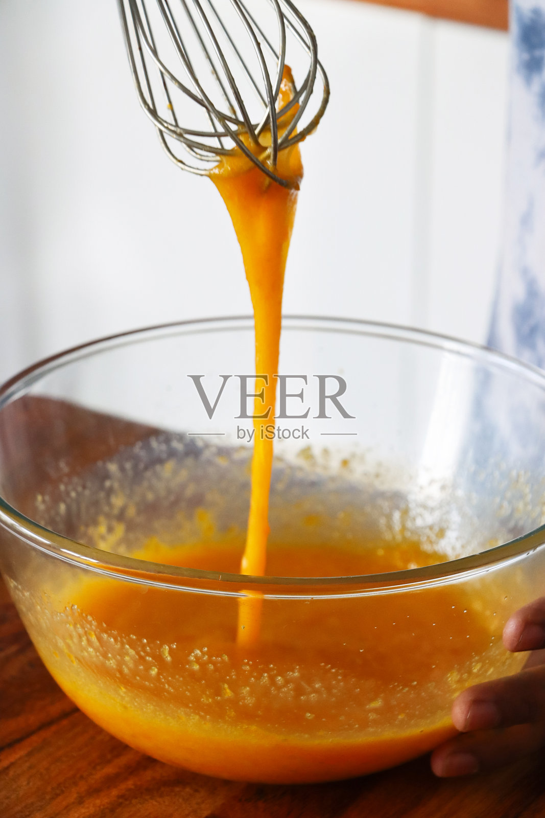 木质厨房台面上的玻璃搅拌碗特写，无法辨认的人正在使用不锈钢打蛋器搅拌鸡蛋和糖，打好的鸡蛋液从打蛋器上滴落，焦点在前景。照片摄影图片