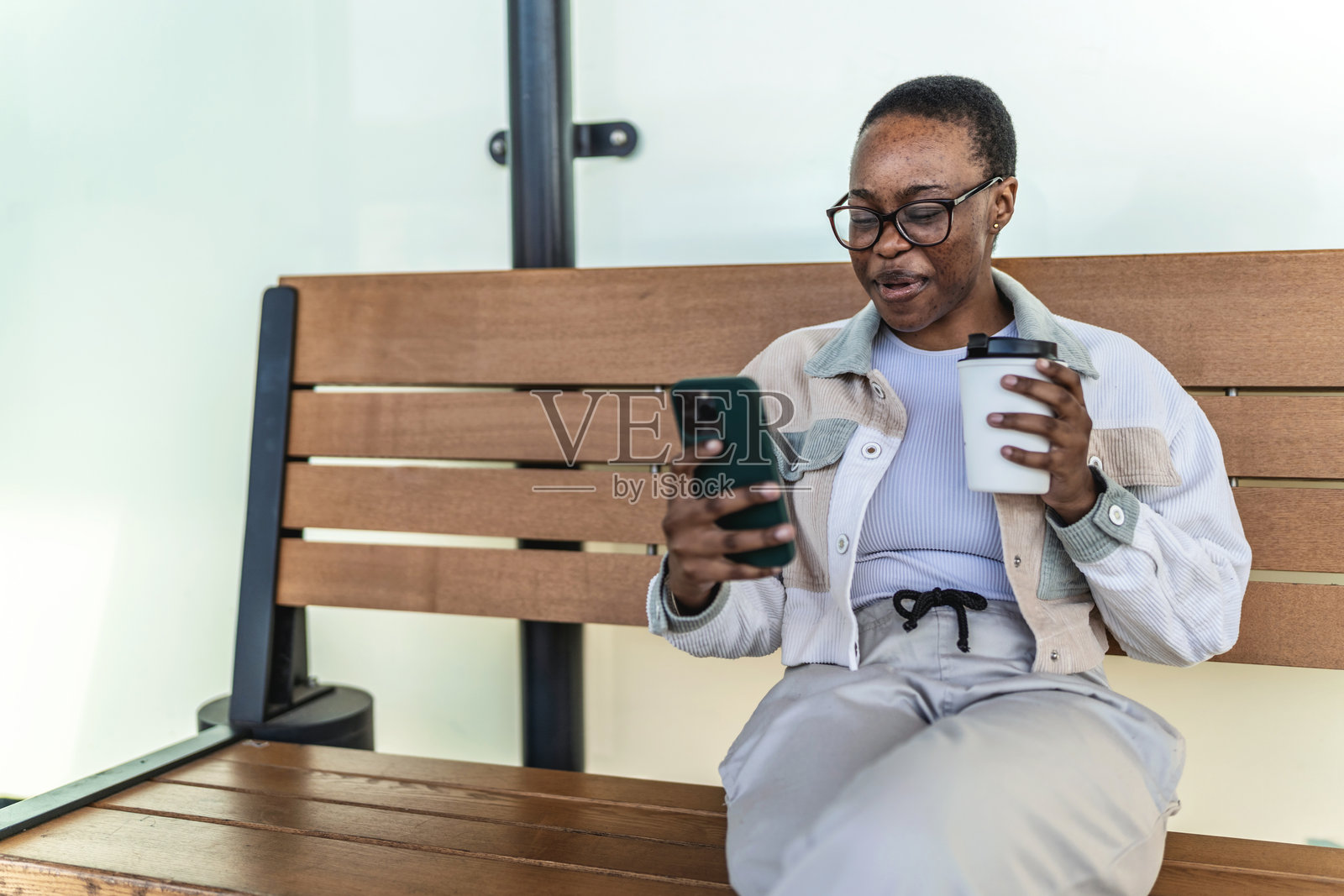 年轻时尚的黑人女性在火车站坐着，手握手机和咖啡，等待地铁列车，同时发短信或观看在线视频。照片摄影图片