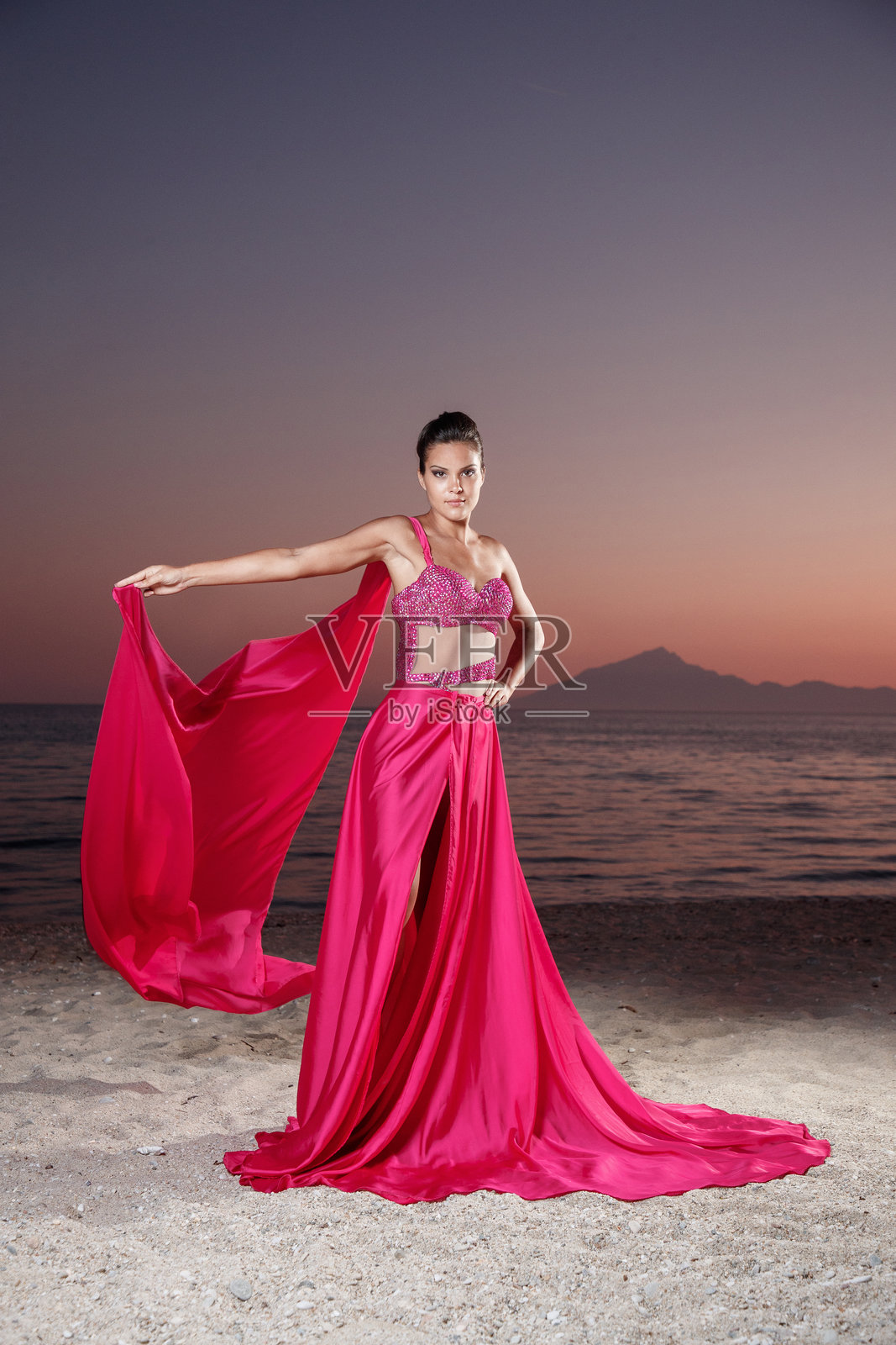 美丽的年轻女子在黄昏的海滩上穿着深红色的面纱和开叉的长裙照片摄影图片