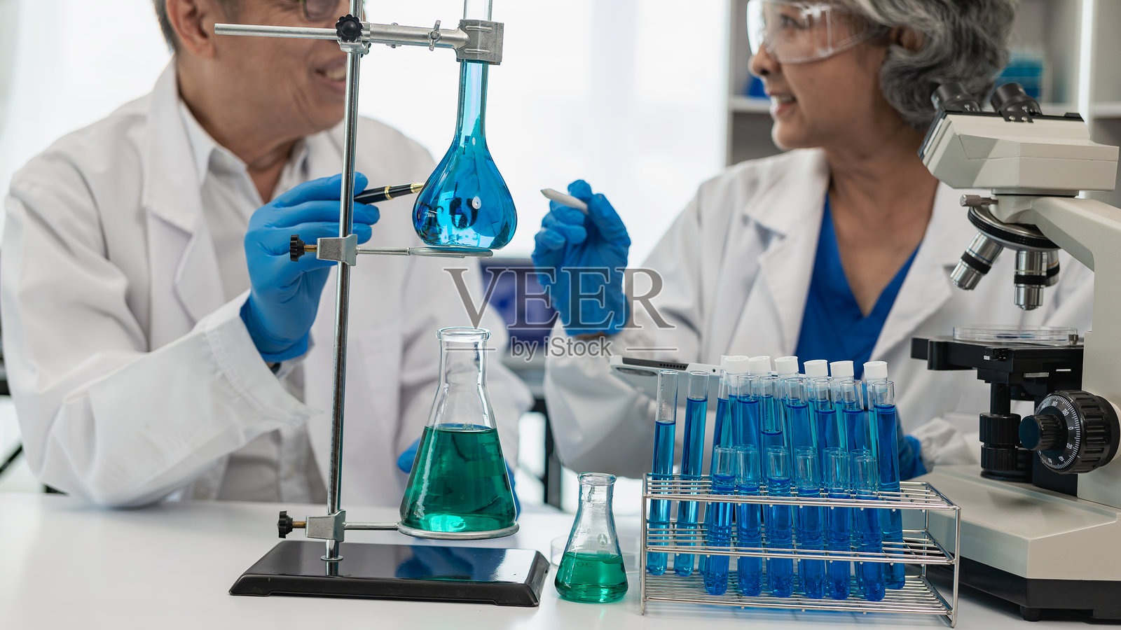 亚洲资深男女化学家在显微镜下检查测试样本。他的助手做了笔记。科学活动在实验室制造疫苗照片摄影图片