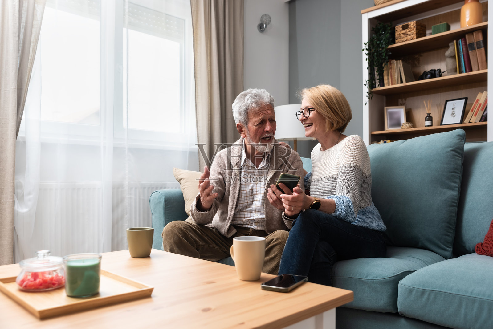 老年夫妇，生活简朴。老年人，男人和女人充满爱和尊重，坐在舒适的家里聊天，在智能手机上看网上有趣的视频，享受他们的退休和生活。照片摄影图片