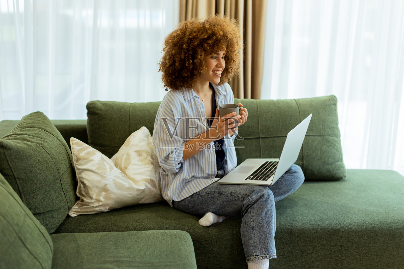 年轻女性在明亮通风的客厅中，享受着温馨的早晨，手里拿着咖啡和笔记本电脑。照片摄影图片
