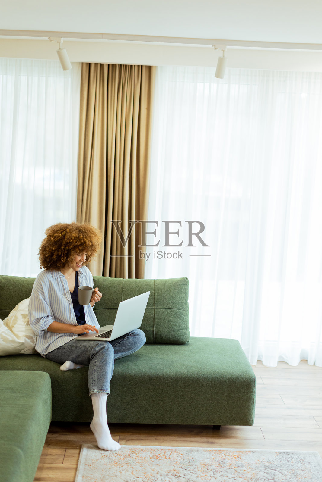 年轻女性在明亮通透的客厅中，享受着温馨的早晨，手里拿着咖啡和笔记本电脑。照片摄影图片