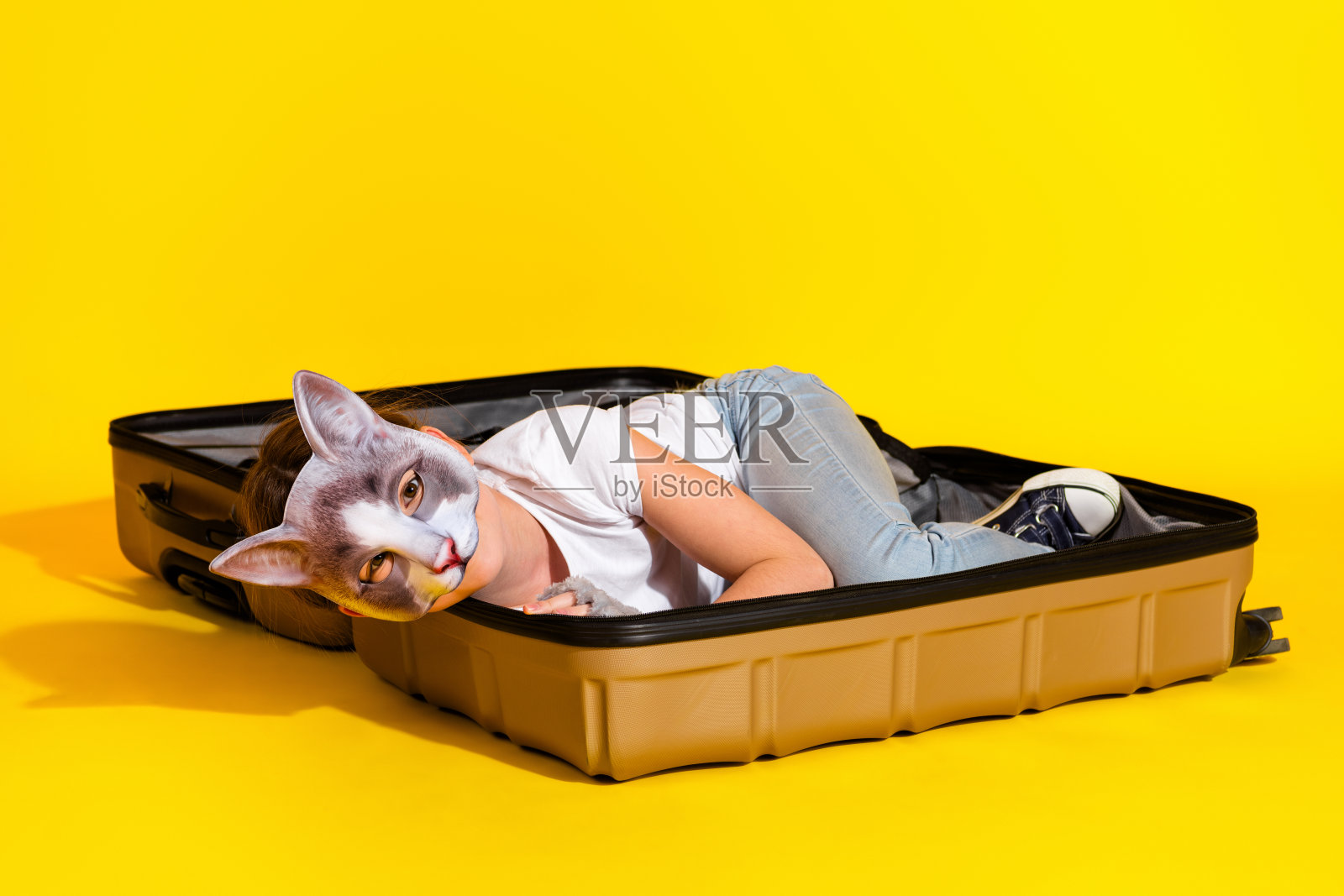 年轻女孩戴着猫面具，躺在一个打开的行李箱里，背景是鲜艳的黄色，象征着创造力和富有想象力的玩耍。照片摄影图片