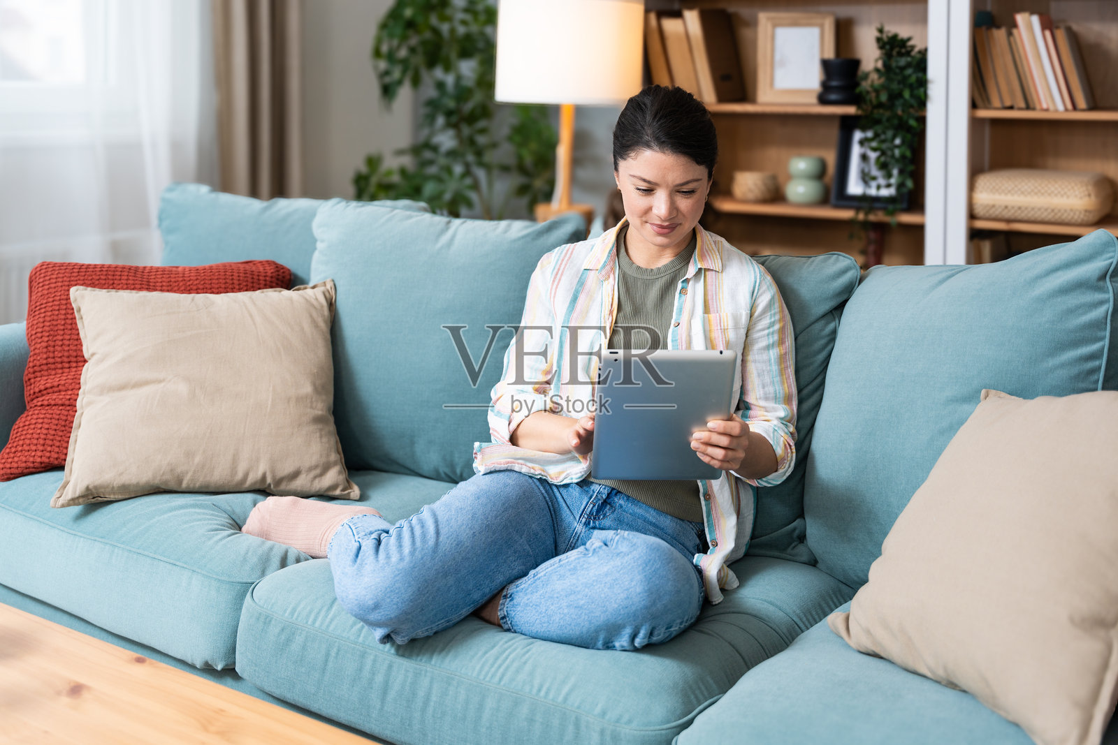 年轻的女性设计专业人士坐在家中的沙发上使用平板电脑。她正在从网上商店选择新的衣橱进行在线购物和订购。照片摄影图片