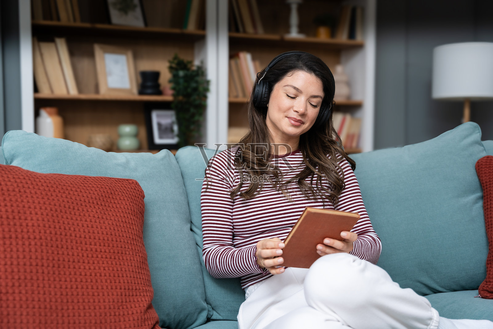 年轻的千禧一代女性坐在家里的沙发上，戴着无线耳机听音乐，享受生活和自由。受过教育的大学生女孩正在休息，放松，听着混音带，阅读书籍。照片摄影图片