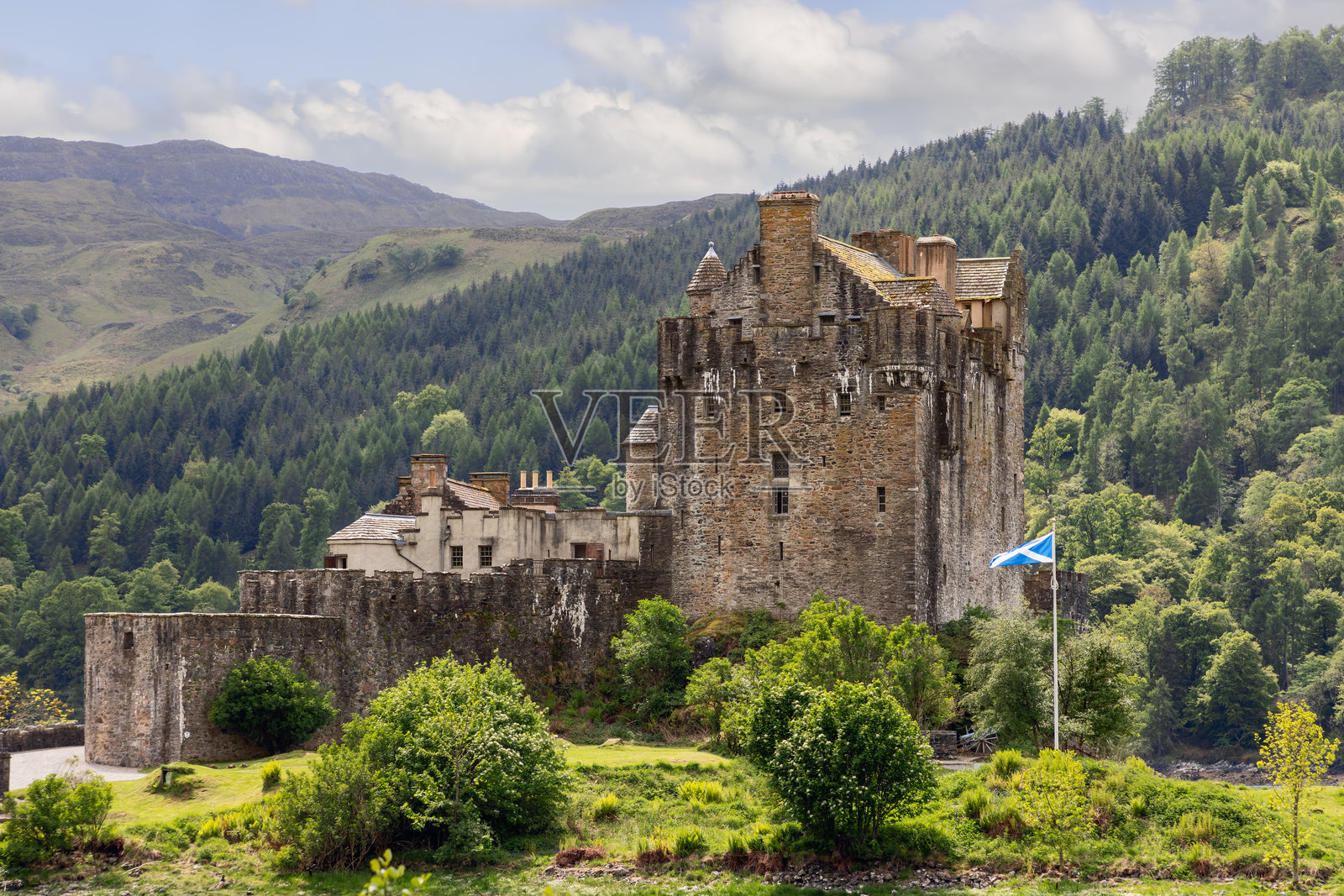 Eilean Donan城堡坐落在郁郁葱葱的绿色植物中，其古老的石头与翠绿的苏格兰高地和飘扬的Saltire国旗形成鲜明对比照片摄影图片