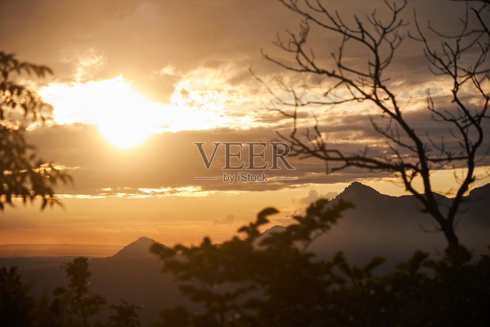 太阳落在山后，在云层和树木的剪影上投下温暖的光芒照片摄影图片