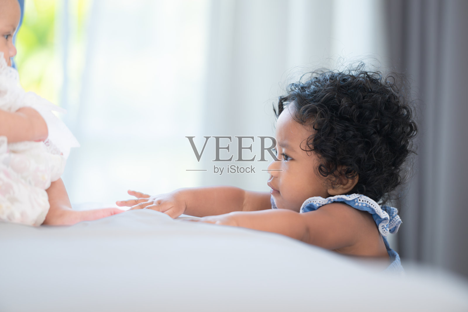 一个小女孩站在床脚，和妈妈抱着的弟弟玩着，脸上带着深情的微笑，卧室里充满了温暖和亲情照片摄影图片