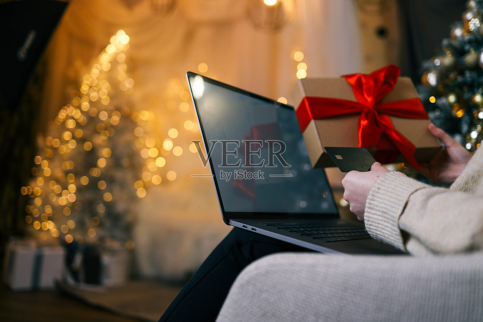 一位女性手持红色丝带的礼品盒和信用卡，正在使用笔记本电脑在线购物，背景中有一棵闪耀的圣诞树，营造出节日和温馨的氛围。照片摄影图片