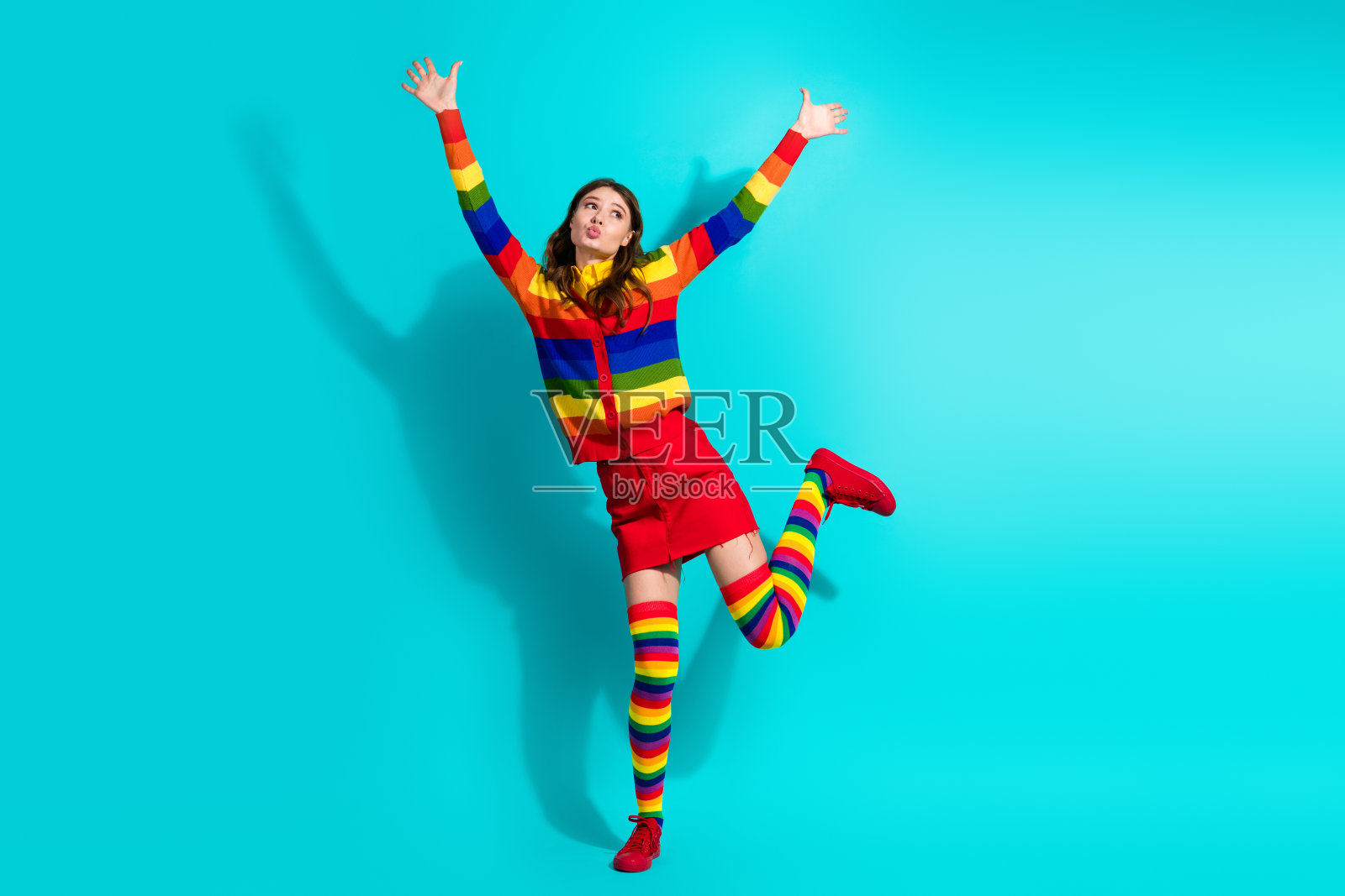 充满活力的年轻女性穿着五彩缤纷的彩虹服装，愉快地在明亮的青绿色背景前跳跃，展现个性、时尚和积极态度。照片摄影图片