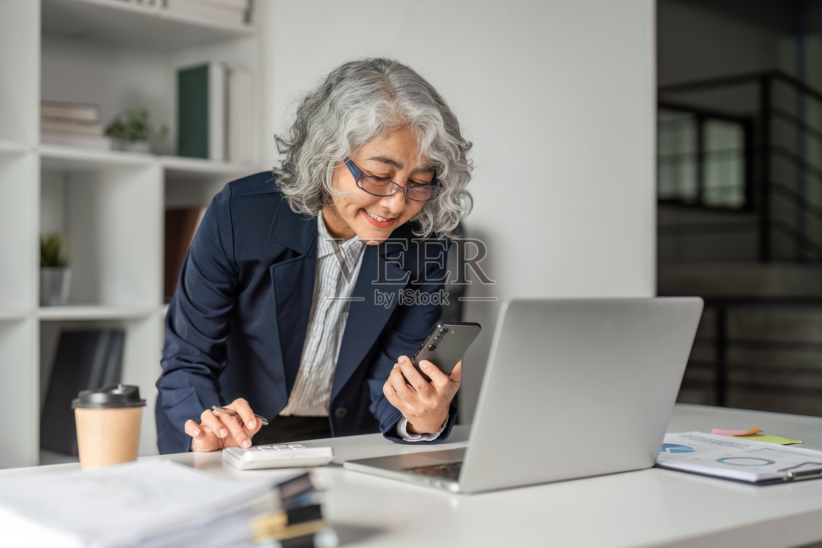 微笑的女性高管在现代办公室使用智能手机并分析笔记本电脑上的报告照片摄影图片