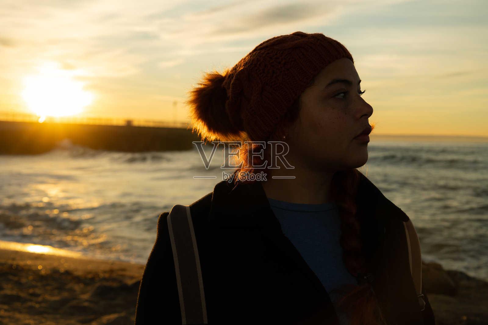背光下的海滩女人，戴着冬帽，夕阳西下。照片摄影图片