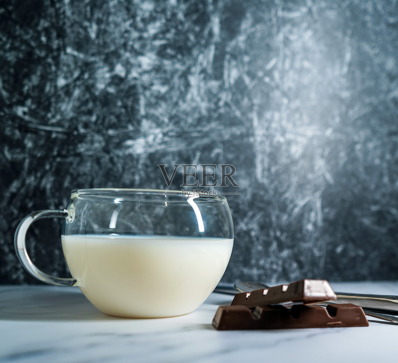一杯牛奶旁边放着两块巧克力，前景中还有一把复古勺子和一把刀。照片摄影图片