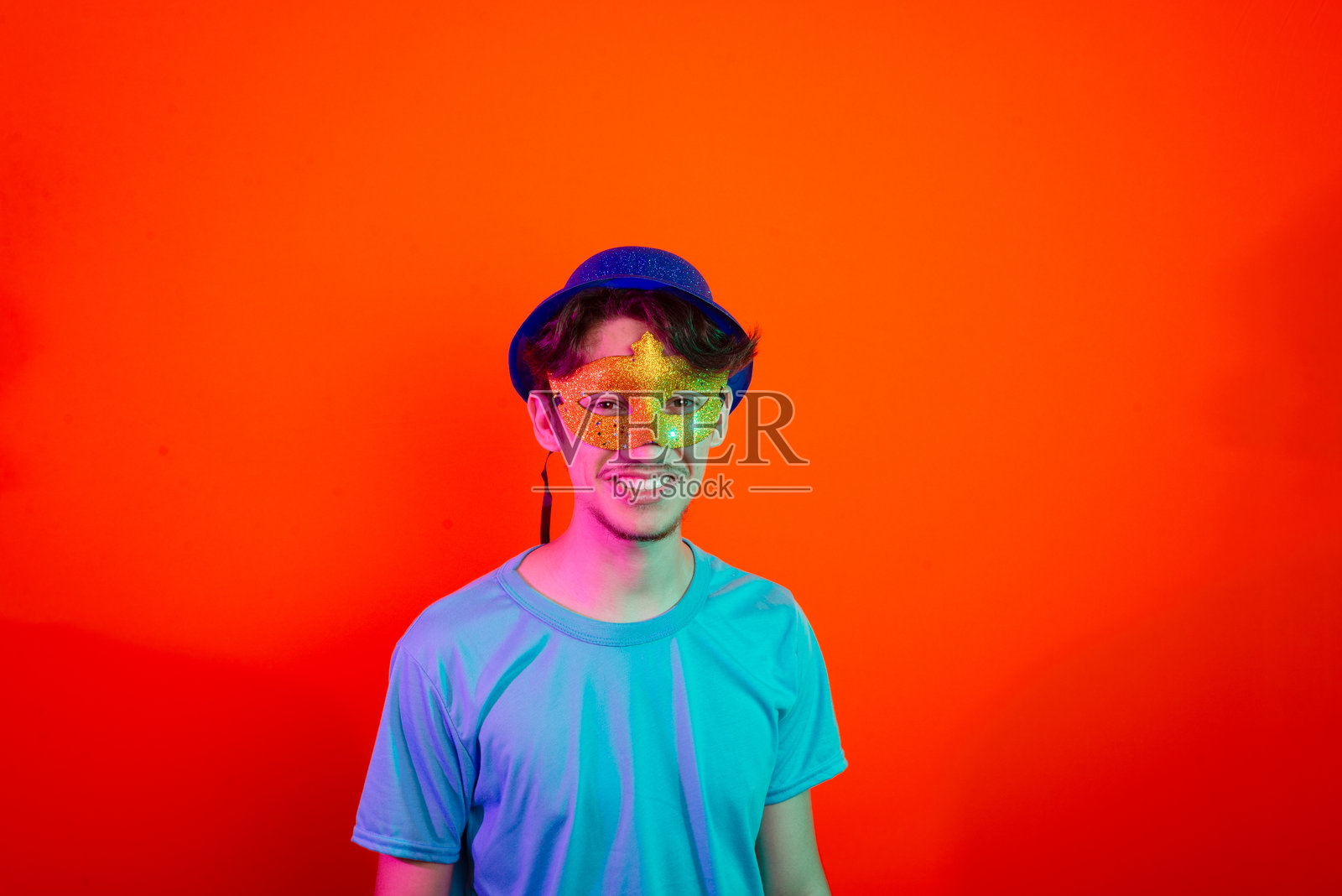 戴着狂欢节面具的帅气年轻男子，面向镜头，快乐而有趣。背景为橙色霓虹灯。照片摄影图片