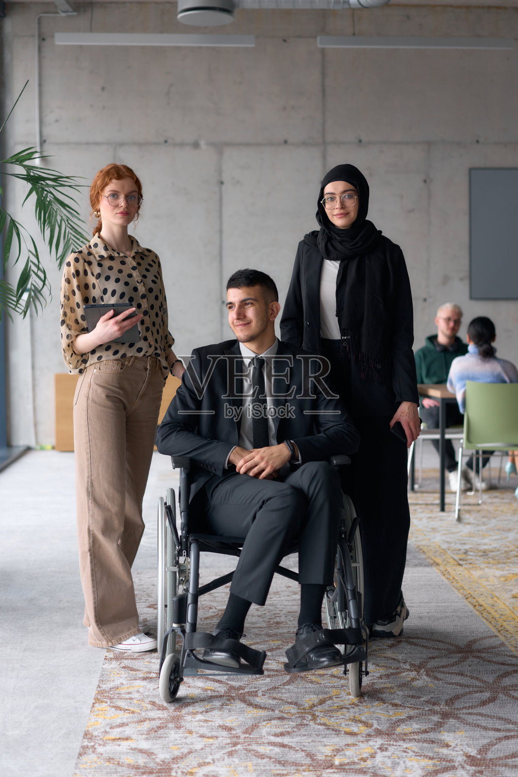 一个多元化的商业团队自信地摆姿势，坐在轮椅上的微笑董事被两位专业女性同事陪伴，一位戴着头巾，另一位手持平板，背景是现代办公室环境。照片摄影图片