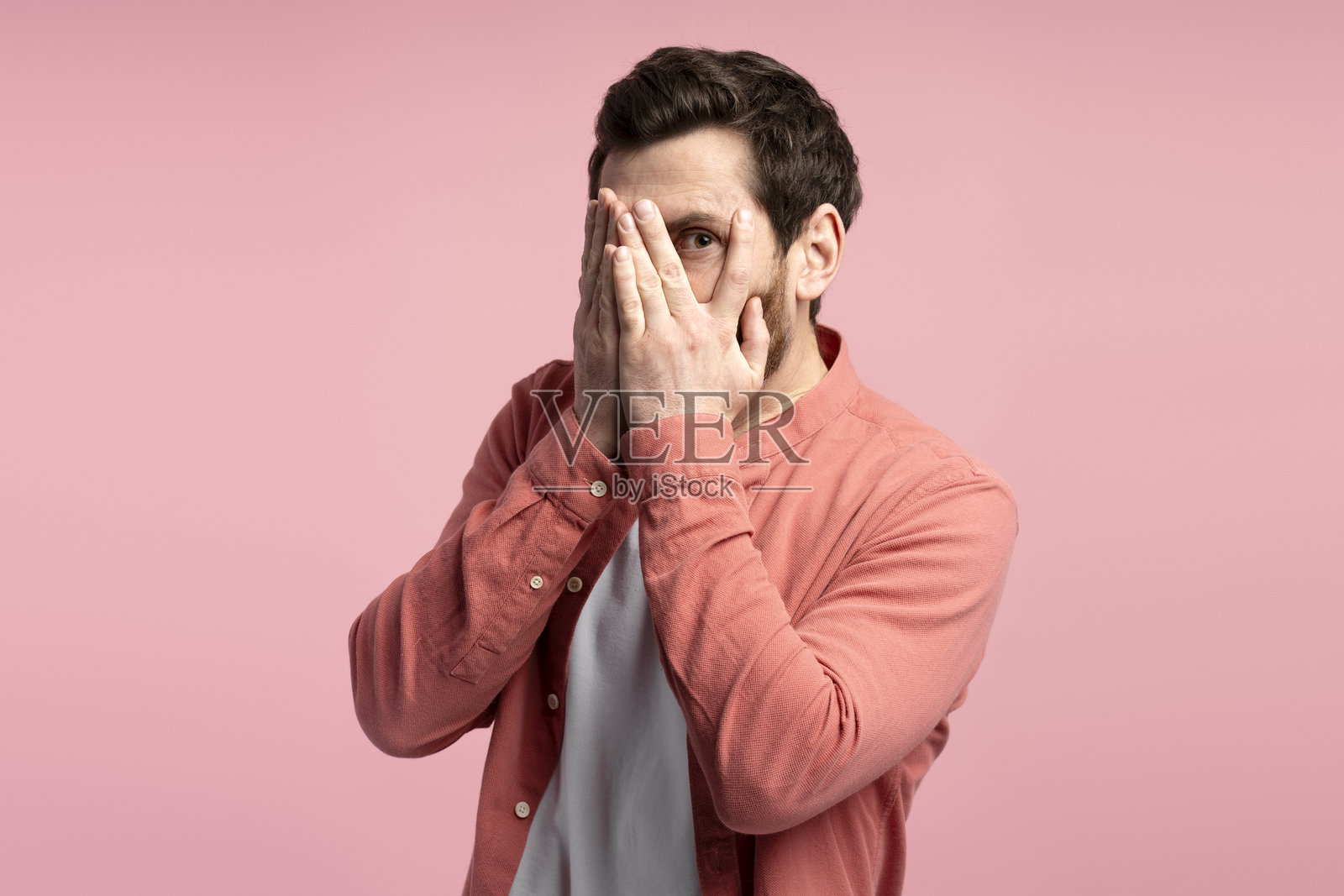 年轻男子穿着衬衫，背景孤立，遮住眼睛并透过手指看。照片摄影图片