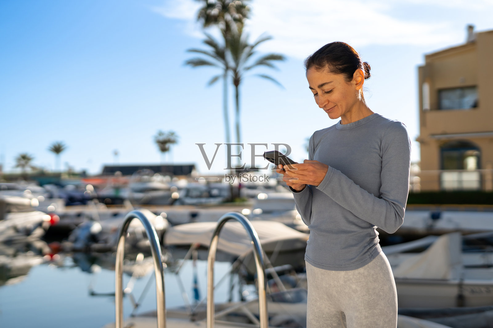 女人在码头附近使用智能手机，周围有船只和棕榈树。照片摄影图片