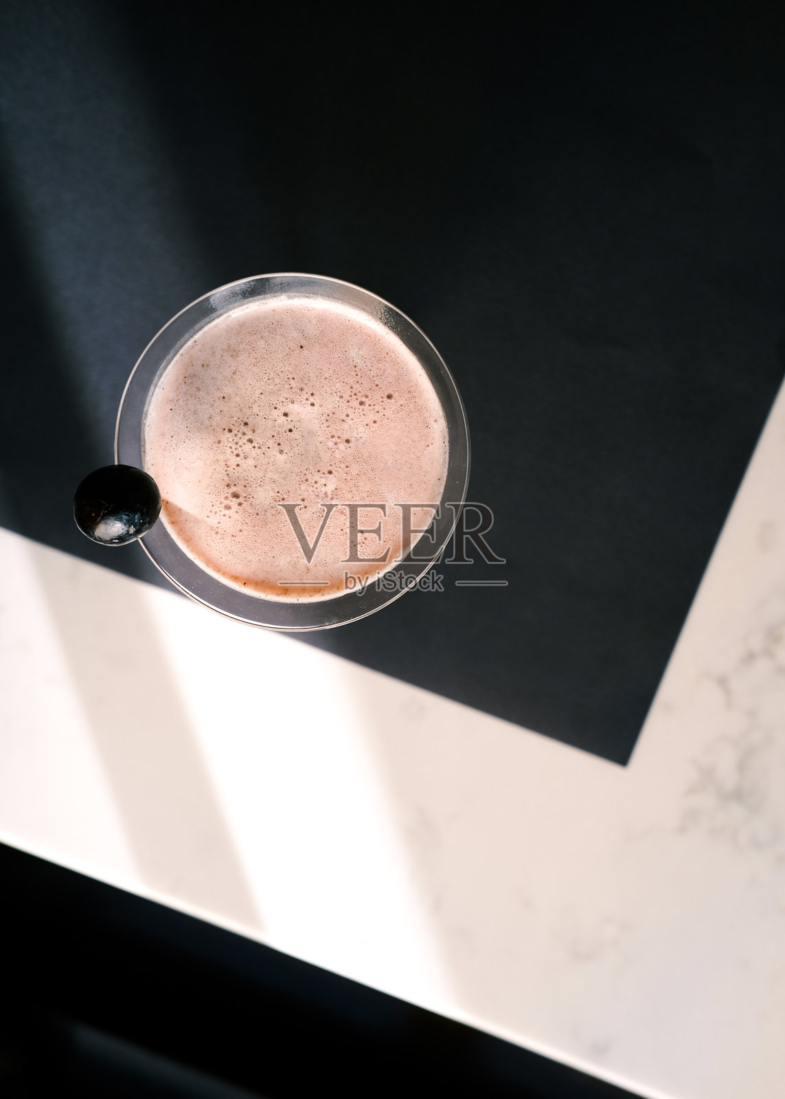 从上方俯视一杯泡沫丰富的粉色鸡尾酒，盛在马提尼酒杯中，点缀着一颗黑橄榄，摆放在现代黑白桌子上，柔和的自然光照射下，优雅的酒吧和奢华的饮品展示。照片摄影图片