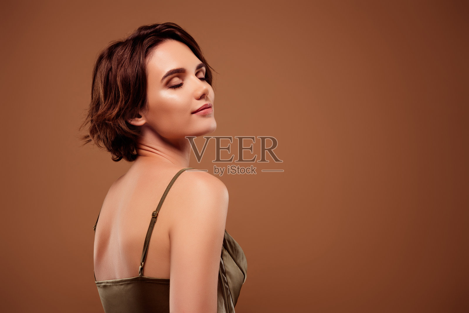 漂亮年轻女孩转身闭眼，穿着时尚的卡其色服装，背景为棕色。照片摄影图片