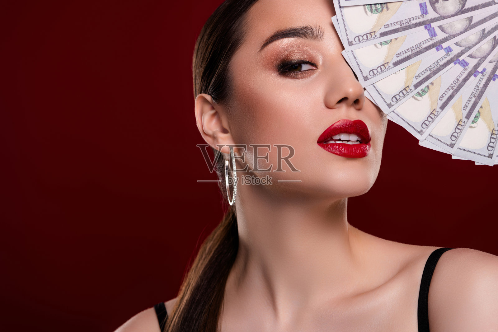特写剪裁的美丽模特女孩手持美元纸币遮住眼睛，背景为深红色。照片摄影图片