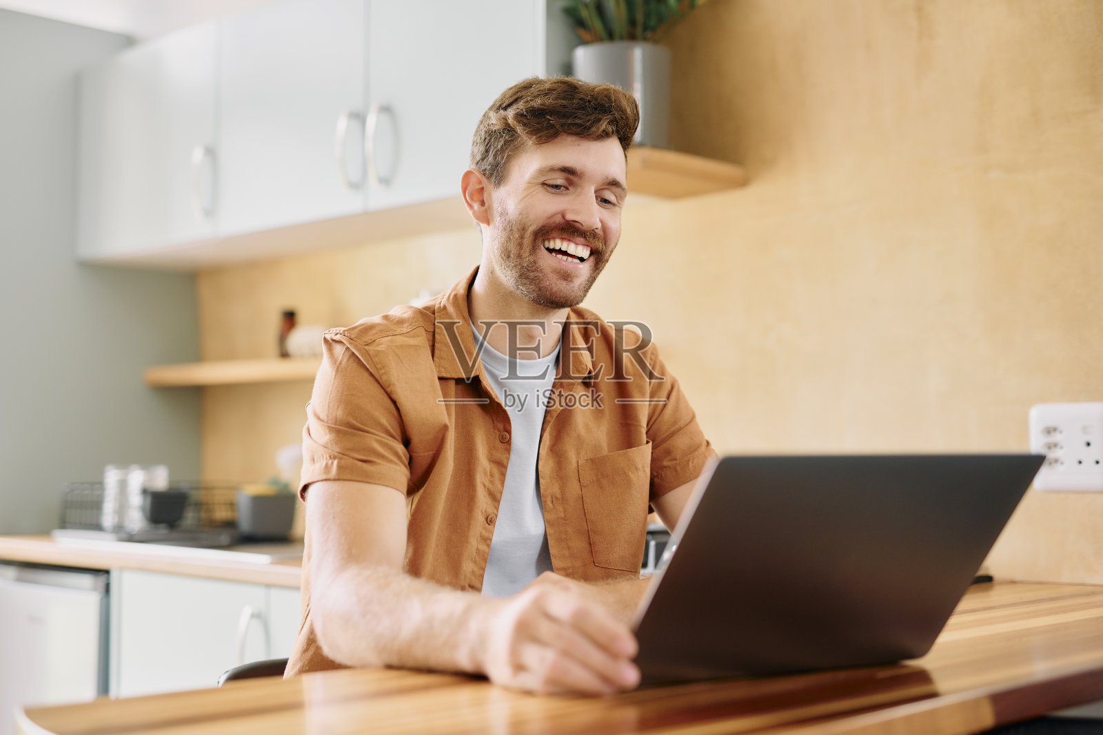 快乐的男人在厨房用笔记本电脑进行视频通话，进行在线交流、交谈或联系。男性，记者或用户，面带微笑使用电脑进行网络更新或聊天应用。照片摄影图片
