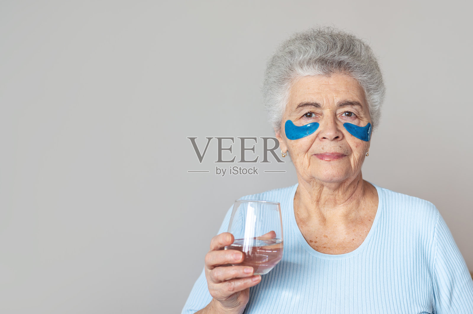 70岁以上的老年女性在家中护理肌肤，眼下有斑点，手中拿着一杯水。宣传美容、自我护理、补水和对衰老的自信。退休人员。留白空间。照片摄影图片