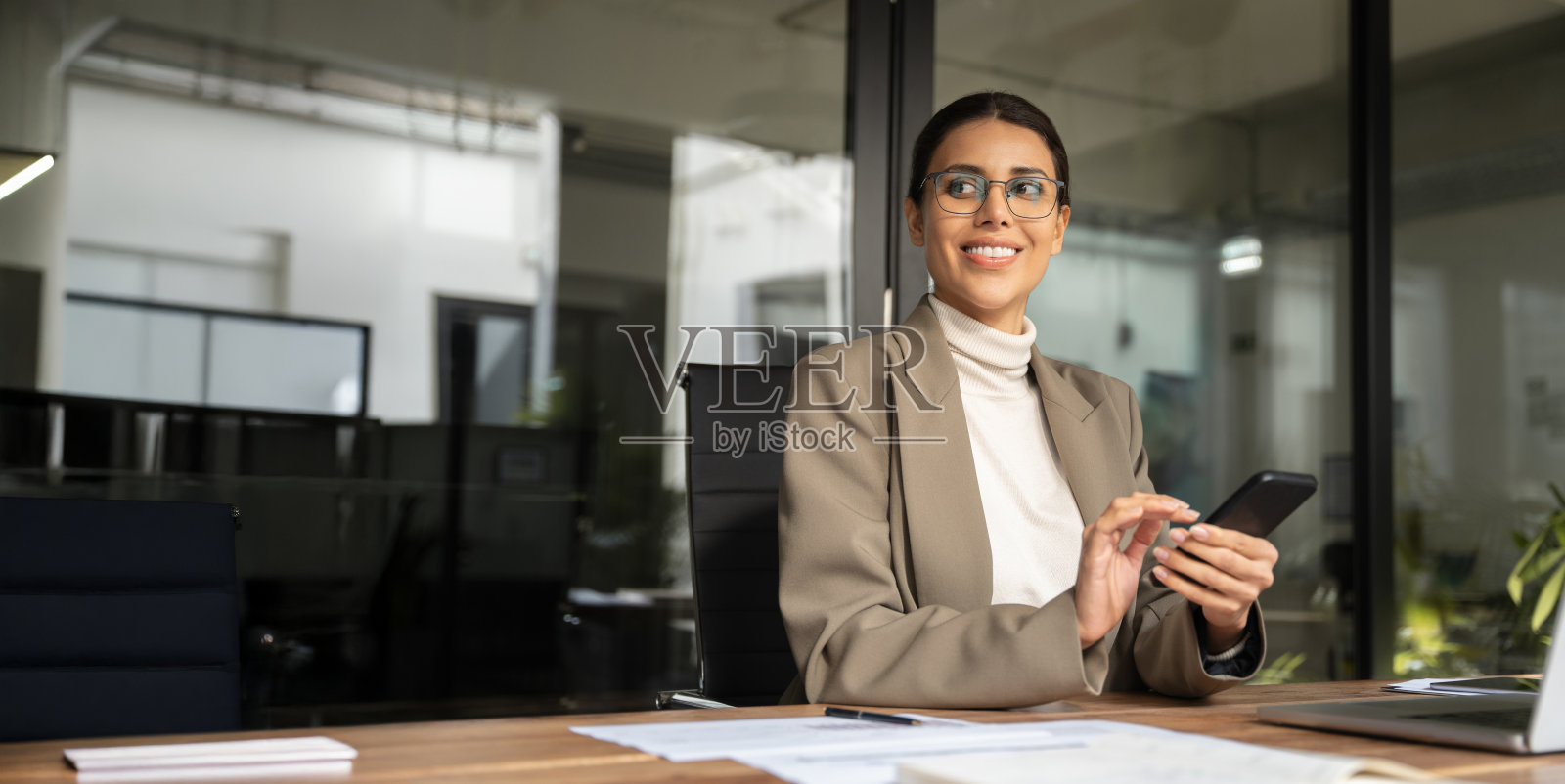 30岁的中东女性商务人士在使用手机应用程序工作，面带微笑的拉丁裔女性IT专家坐在现代办公室的桌子旁，手持智能手机。留白空间。照片摄影图片