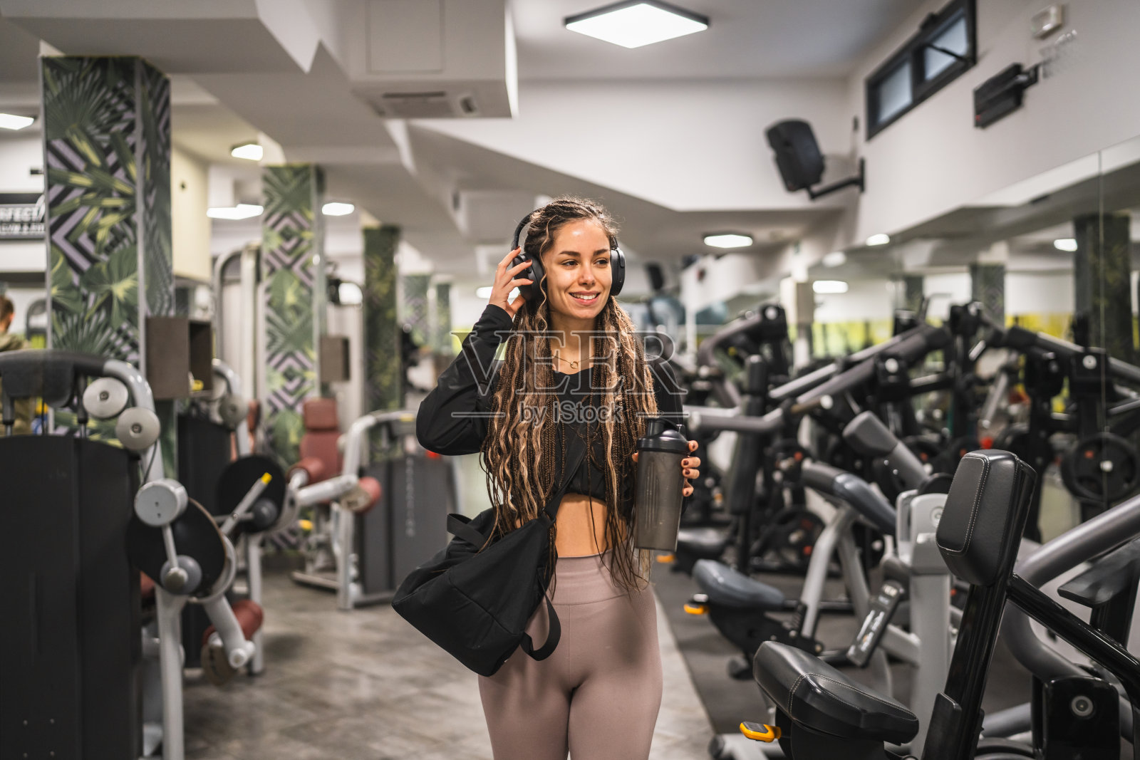 年轻女性戴着耳机，手持摇摇杯走过健身房照片摄影图片