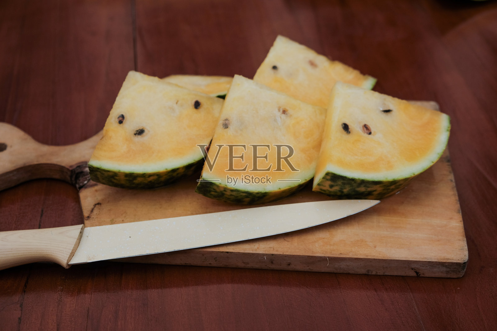 一片带籽的黄色西瓜三角形切片，旁边放着一把厨房刀，放在木质砧板上。照片摄影图片