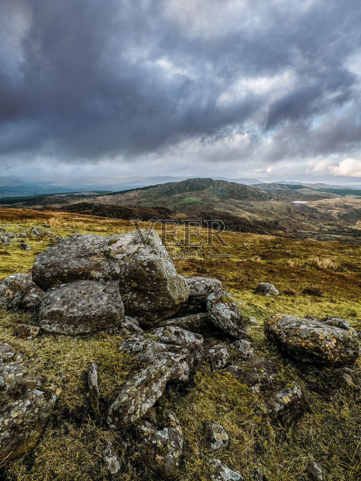 一幅风景画，山坡上散布着巨石，远处是起伏的绿色山丘。天空阴云密布，为地形营造出戏剧性的氛围。照片摄影图片