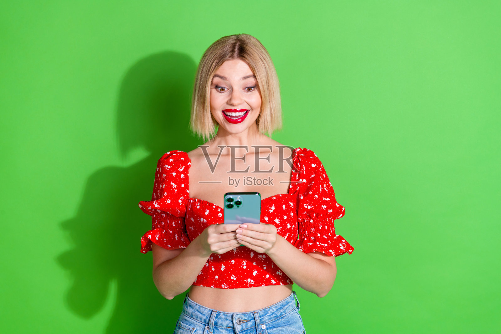 穿着红色时尚服装的可爱女孩手持现代设备，背景为绿色。照片摄影图片