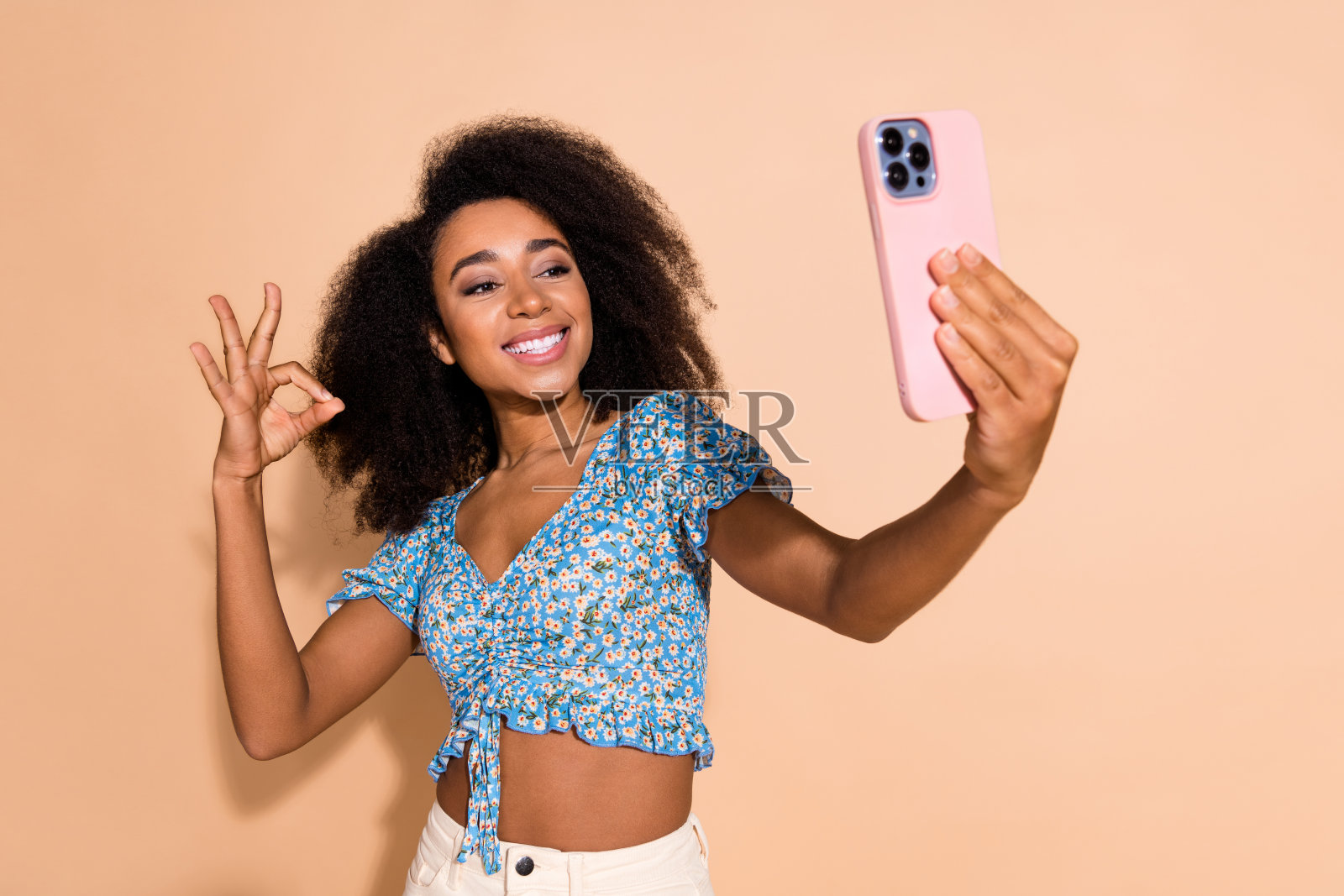 可爱的女孩穿着蓝色上衣，正在用现代设备录制视频，做出“OK”手势，背景为米色。照片摄影图片