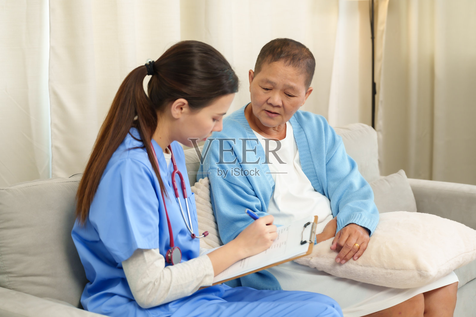 年轻护士坐在沙发上与正在家中康复的老年亚洲女性进行咨询。护士手持夹板写笔记，同时在康复期间进行支持性的医疗对话。照片摄影图片