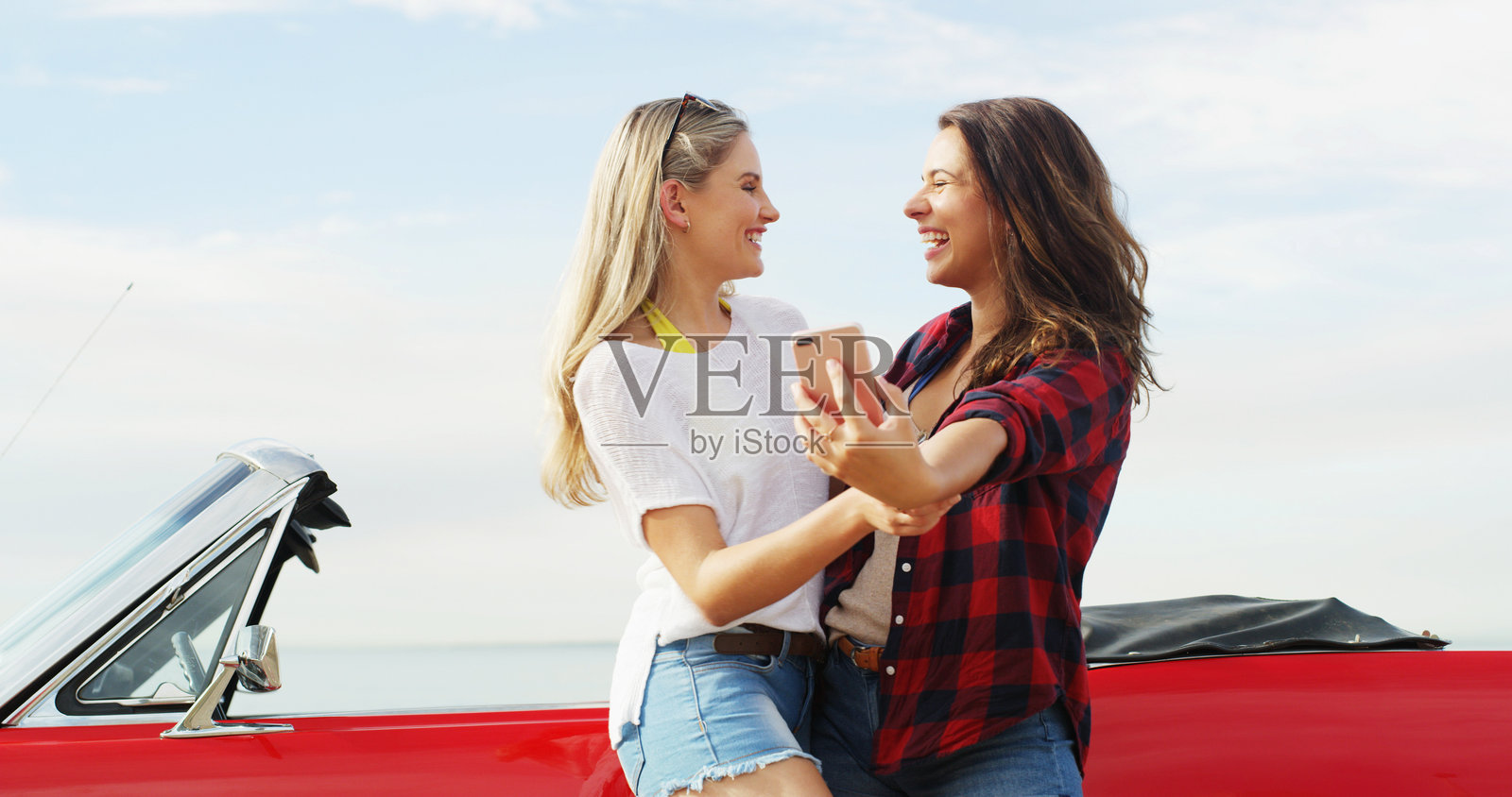 公路旅行，朋友们在车旁欢笑并自拍，适合旅行、度假或户外活动。敞篷车，快乐的女性，记录旅途中的有趣回忆或搞笑的社交媒体帖子。照片摄影图片