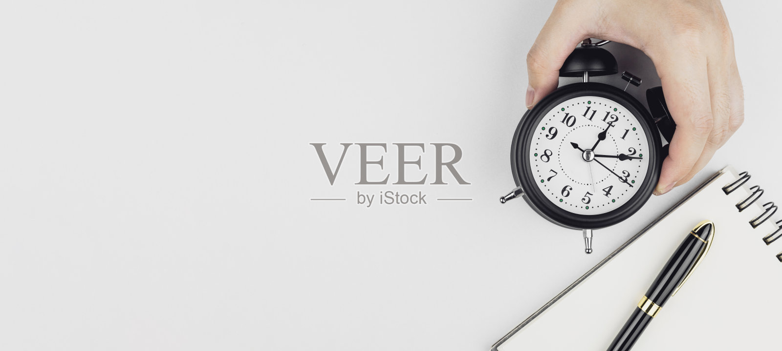 手握黑色闹钟，旁边有笔记本和钢笔，背景为白色。照片摄影图片