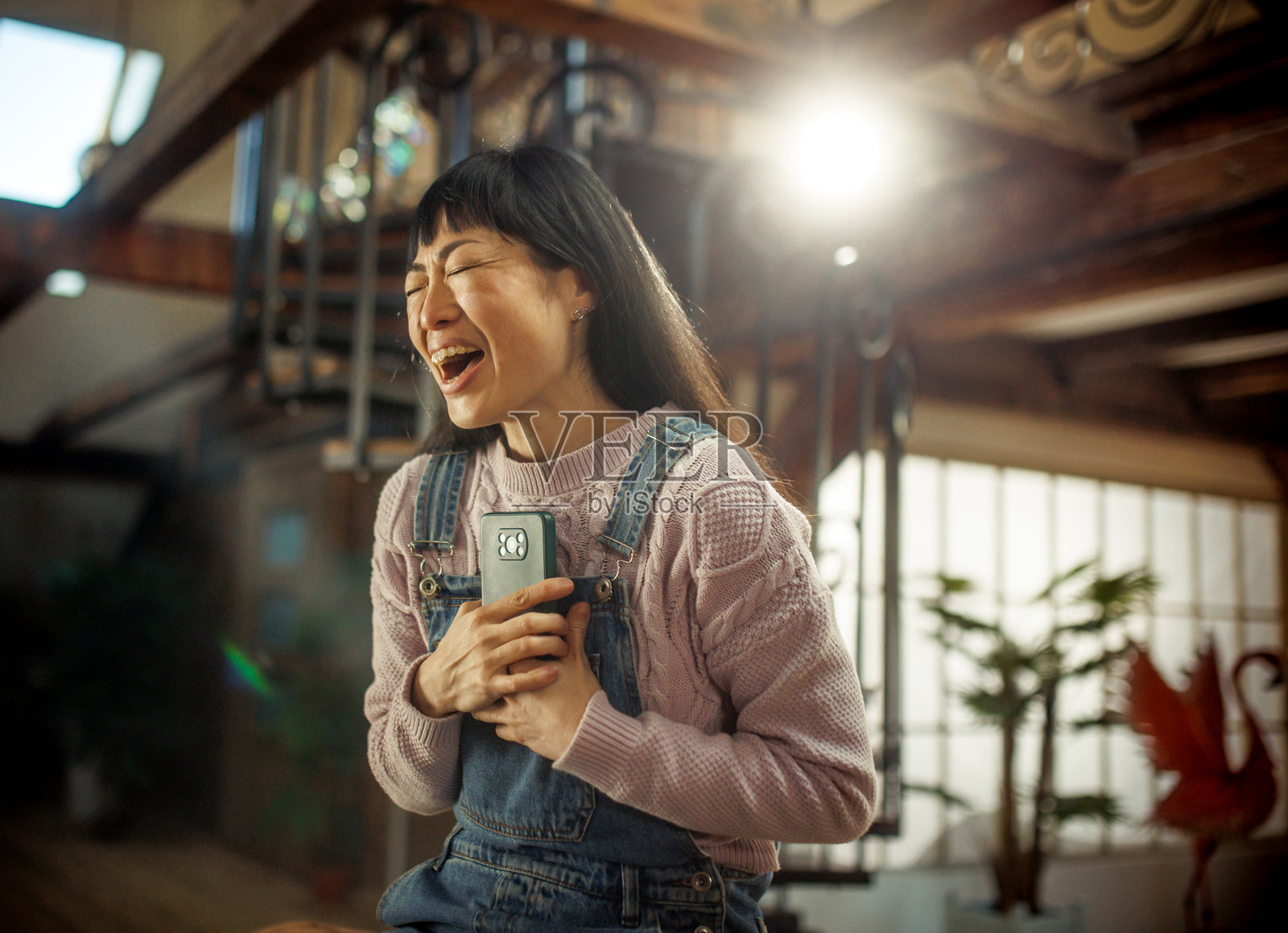 快乐的年轻千禧一代日本女性坐在家里的沙发上使用手机。美丽的女性在看手机屏幕时笑得很开心。科技与社交媒体概念。照片摄影图片