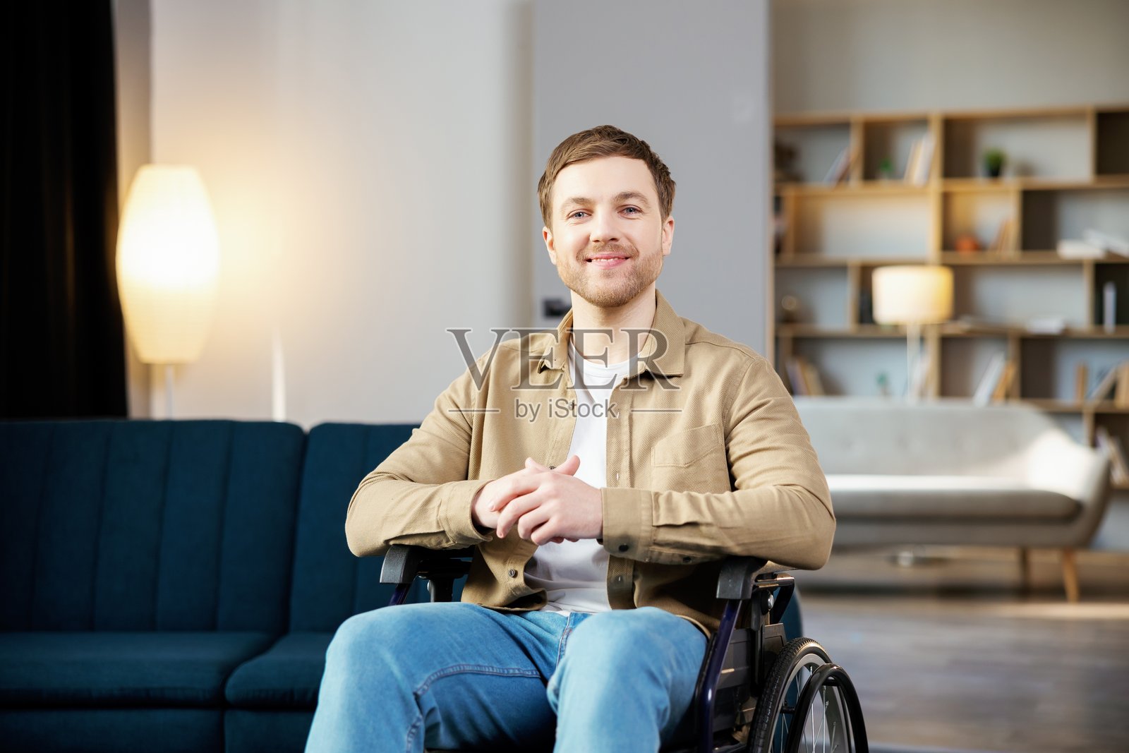 积极的生活方式，关心残疾人，在家中进行康复和支持。年轻的白人男性坐在轮椅上，微笑着看向镜头，身处客厅。照片摄影图片
