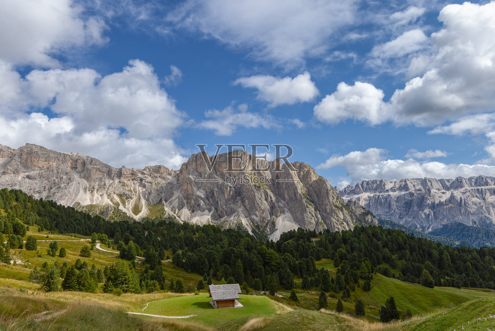 意大利多洛米蒂山的田园风光，木屋被草地和针叶林环绕。高耸的石灰岩山峰在明亮的蓝天和蓬松的白云下矗立。照片摄影图片