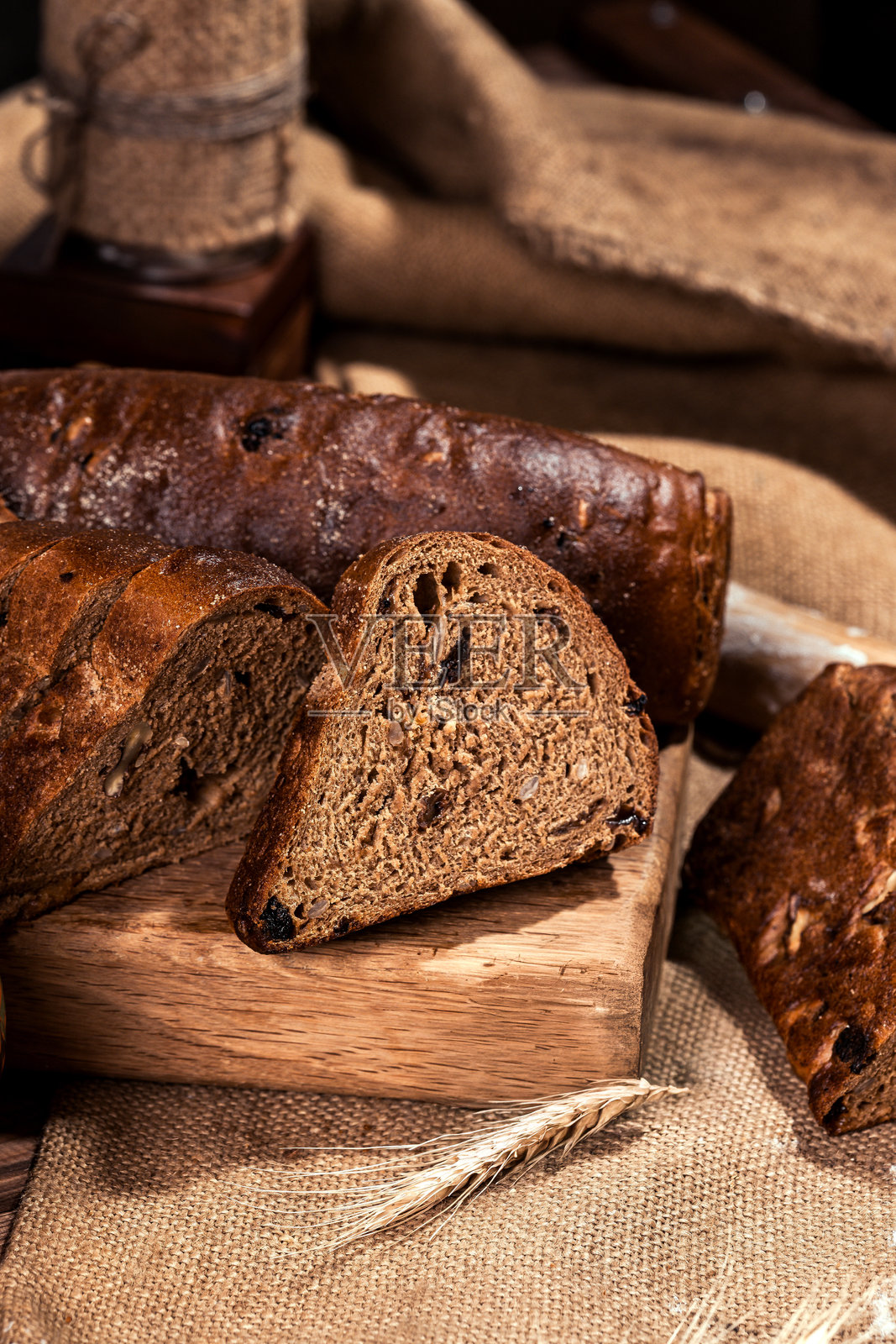 含有葡萄干、干杏、向日葵种子、孜然种子的黑麦面包，两个面包放在木质背景上，面粉、筛子、擀面杖、鸡蛋，自制面包，小麦穗，涂抹黄油和牛奶的黑面包照片摄影图片