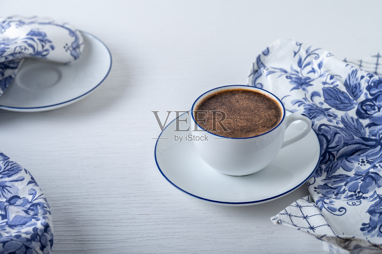 白色咖啡杯配碟子，蓝白相间的餐巾放在白色桌子上。照片摄影图片