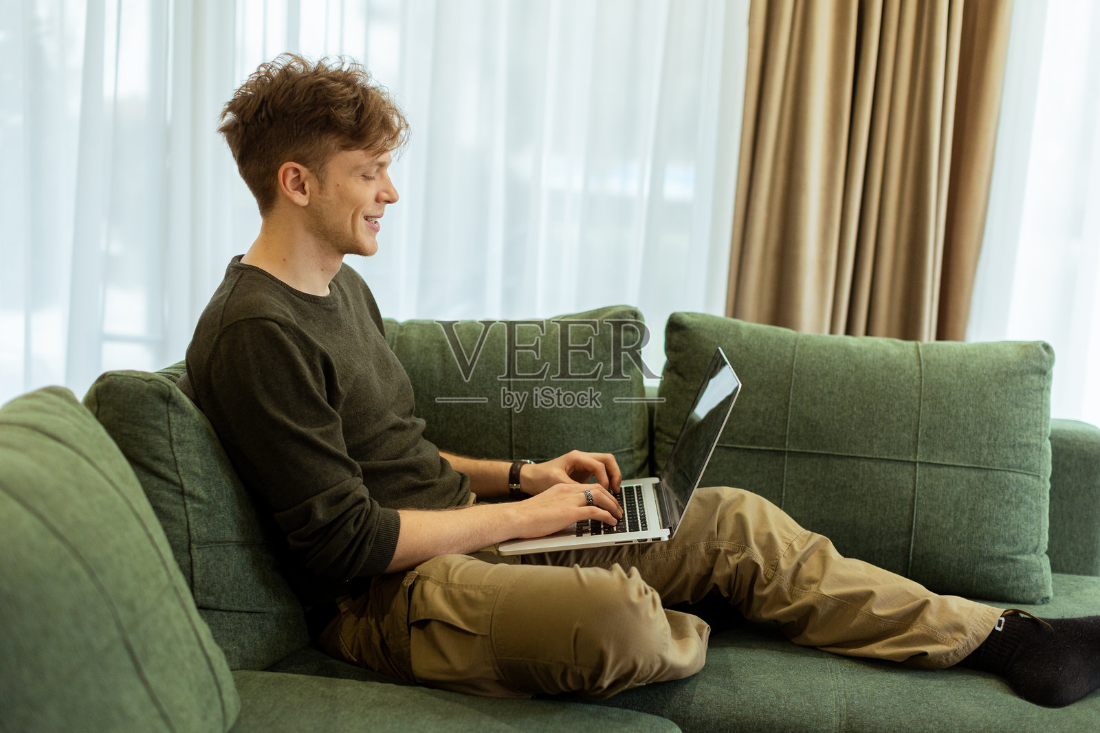 年轻男子在舒适的客厅里，放松地坐在绿色沙发上，专注地使用笔记本电脑，阳光透过窗户洒入室内。照片摄影图片