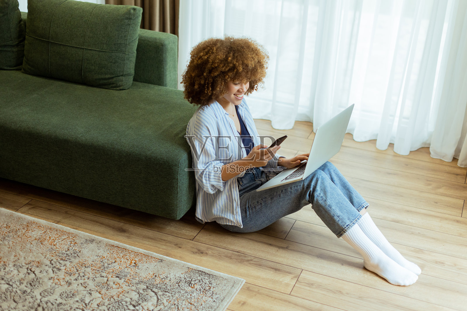 年轻女性在明亮舒适的客厅地板上放松，手里拿着笔记本电脑和智能手机，和谐地平衡着工作与休闲的下午时光。照片摄影图片