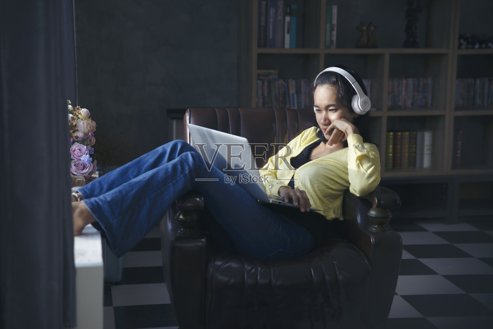 亚洲女性坐在沙发上，戴着耳机，愉快地听着笔记本电脑里的音乐，享受假期时光。照片摄影图片