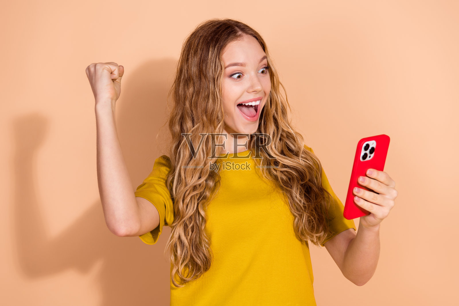 兴奋的年轻女性，卷发庆祝着，手中拿着一部鲜艳的红色智能手机，背景为米色。照片摄影图片
