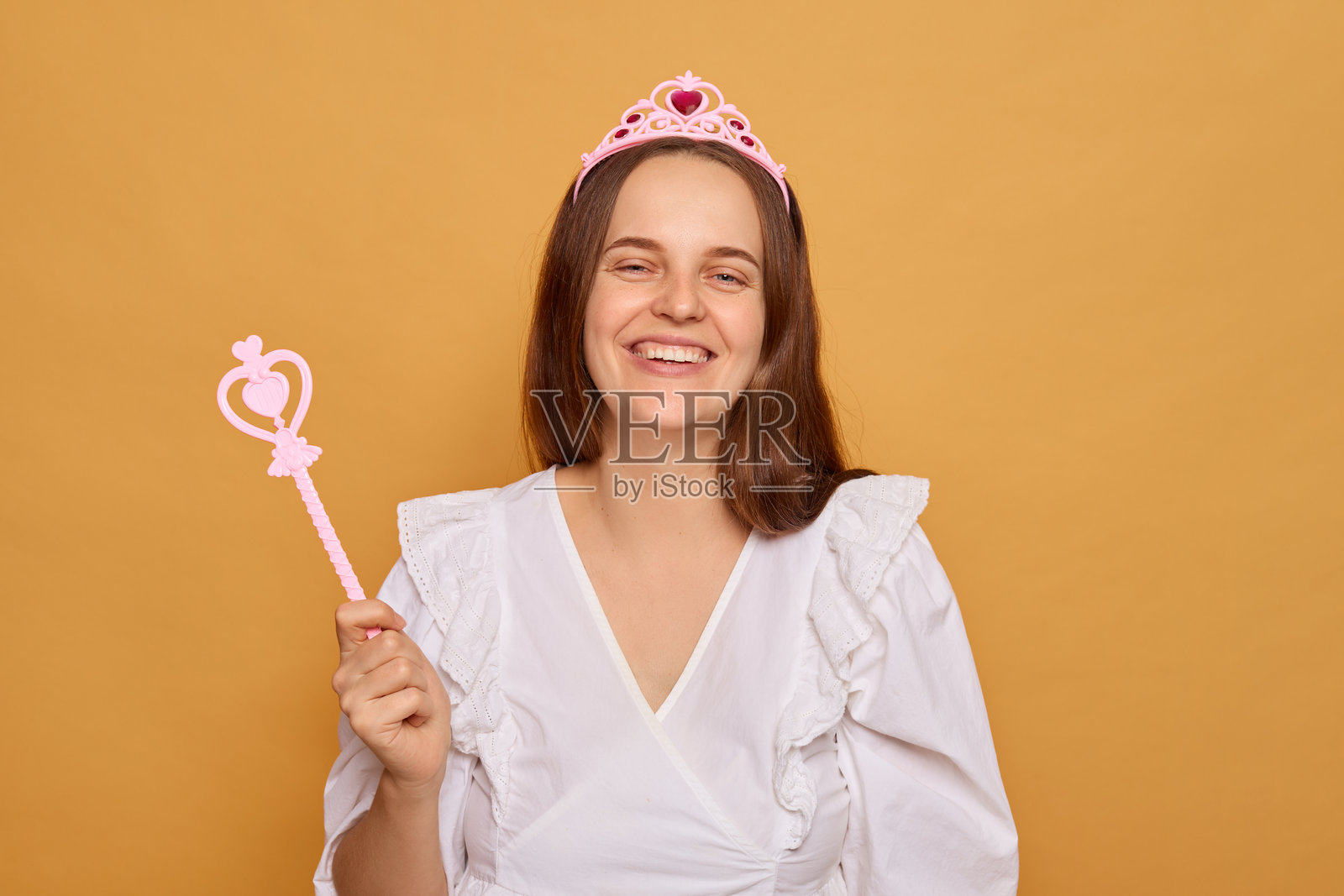一位女性穿着白色连衣裙和粉色王冠，灿烂地微笑。她手持粉色魔杖，在明亮的黄色背景前散发出俏皮而神奇的气息。照片摄影图片