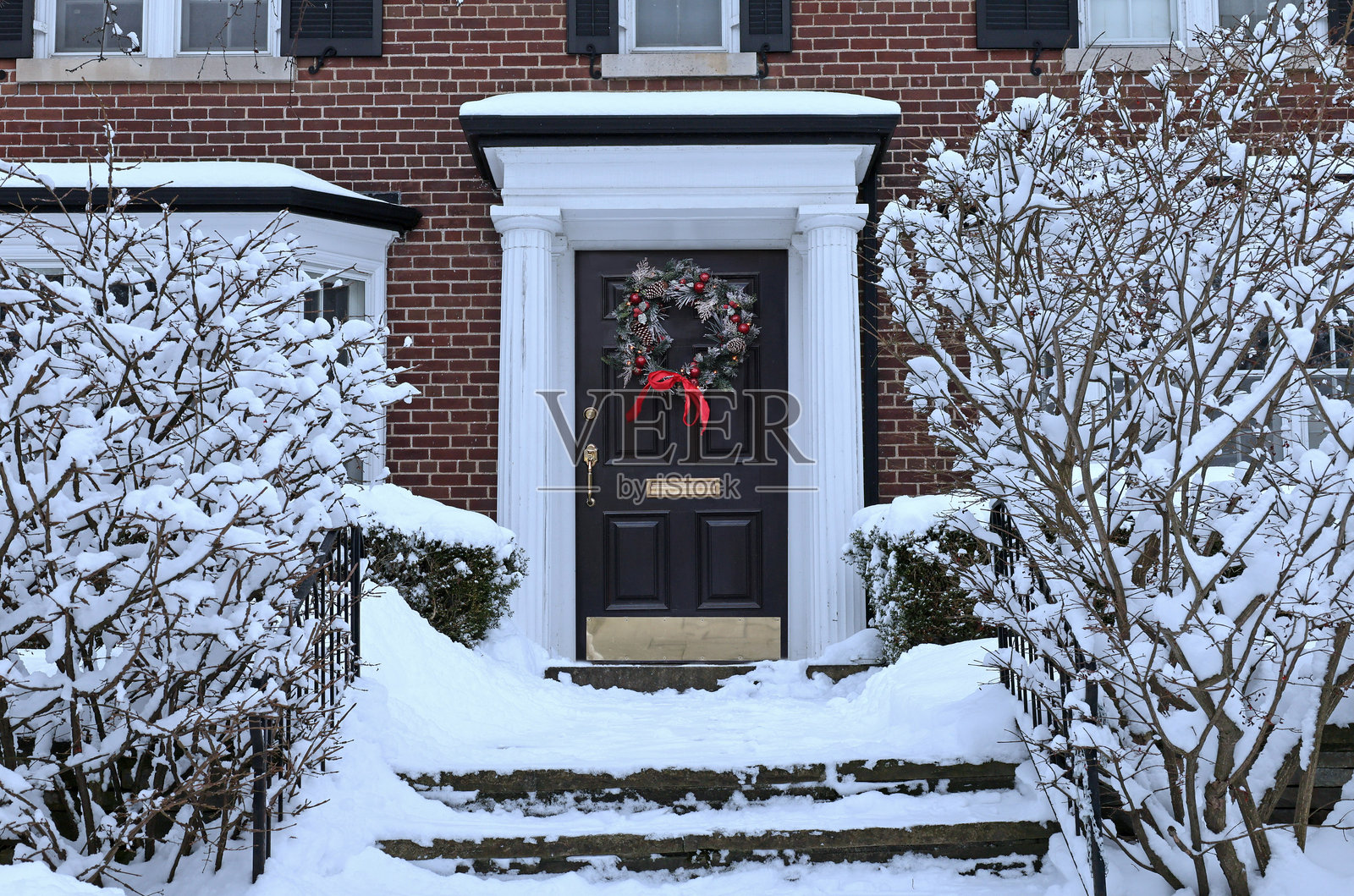 传统的郊区住宅，灌木丛覆盖着雪，前门挂着装饰花环照片摄影图片
