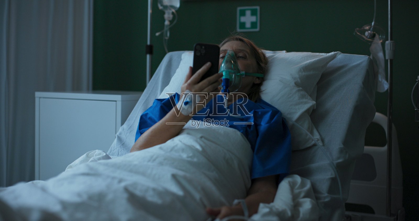 医院病房中的老年女性，戴着氧气面罩，正在玩手机游戏以消遣。照片摄影图片