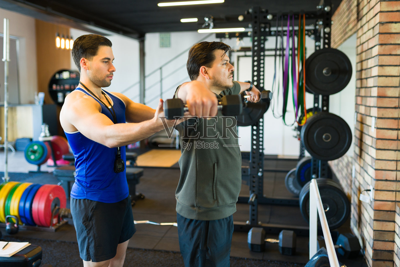 专注的男性客户在现代健身房中举哑铃，私人教练在旁协助进行力量训练。照片摄影图片